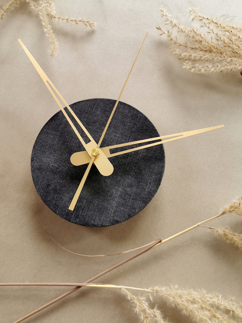 May include: A black clock with gold hands on a beige background. The clock face is round and has a textured surface.