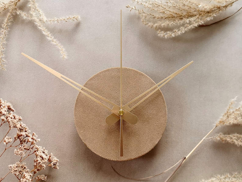 May include: A round, beige clock with gold hands. The clock is on a light gray background with dried flowers around it.