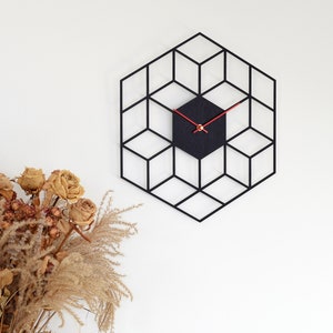 May include: A black geometric clock with red hands on a white wall. The clock is shaped like a hexagon and has a 3D effect. There are dried flowers in a vase in the bottom left corner of the image.