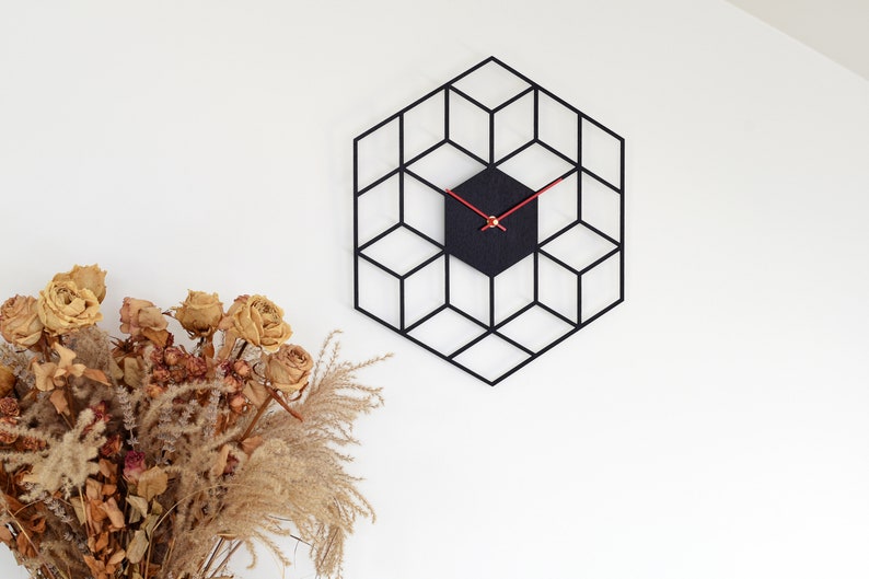 May include: A black geometric clock with red hands on a white wall. The clock is shaped like a hexagon and has a 3D effect. There are dried flowers in a vase in the bottom left corner of the image.