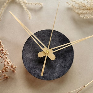May include: A black clock with gold hands on a beige background. The clock face is made of a textured fabric.