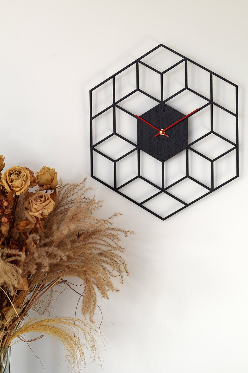 May include: A black geometric wall clock with red hands. The clock is in the shape of a hexagon and has a 3D effect. The clock is hanging on a white wall.