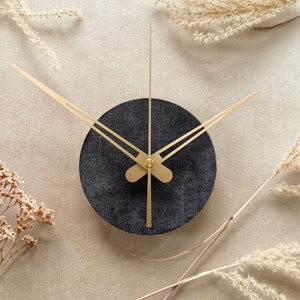 May include: A black clock with gold hands against a beige background. The clock is surrounded by dried flowers.