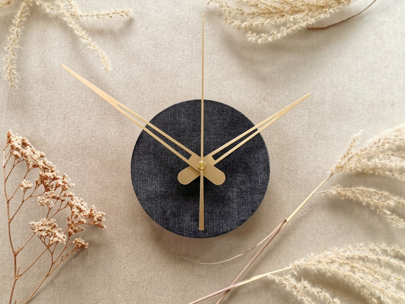 May include: A black clock with gold hands against a beige background. The clock is surrounded by dried flowers.