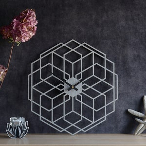 May include: A silver geometric wall clock with black hands and a gold centre. The clock is hanging on a dark grey wall with dried flowers in a vase and a decorative candle holder on a wooden shelf.