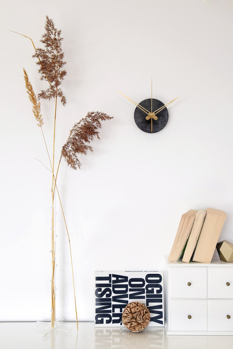 May include: A black and gold clock with thin gold hands on a white wall. A dried grass arrangement is in a clear vase on a white surface. A white dresser with four drawers has a stack of books and a brown decorative object on top. The book is titled "Ogilvy on Advertising".