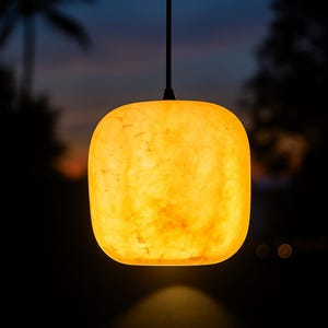 Könnte beinhalten: Eine leuchtende, quadratische Pendelleuchte mit abgerundeten Ecken und einer warmen, bernsteinfarbenen, marmorierten Textur. Die Lampe hängt an einem dünnen, dunklen Kabel und strahlt ein weiches Licht aus, das den Bereich darunter erhellt.