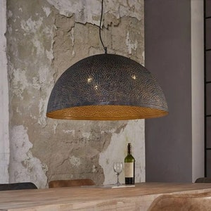 Op de afbeelding: Een zwarte metalen hanglamp met een ronde, geperforeerde kap. De kap heeft een gouden binnenkant en hangt aan een ketting. De lamp hangt boven een houten tafel met een fles wijn en een wijnglas op de tafel.