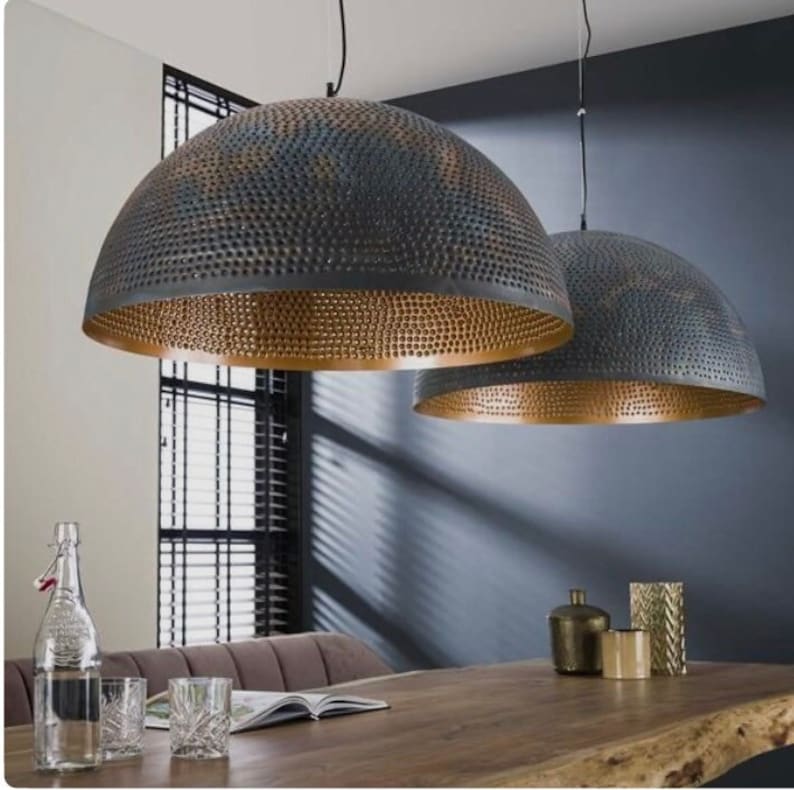Op de afbeelding: Twee grote, ronde hanglampen van metaal met een gehamerde afwerking en een gouden binnenkant. De lampen hangen boven een houten tafel.