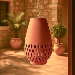 Puede incluir: Una lámpara colgante de color terracota, con forma de lágrima y un patrón calado decorativo. La lámpara está suspendida por una cadena dorada. El fondo presenta plantas en macetas y una pared de piedra, sugiriendo un entorno exterior.