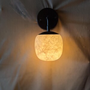 May include: A wall-mounted light fixture with a black circular base and curved arm supporting a textured, off-white globe-shaped shade. The shade emits a warm, inviting glow. The fixture is designed for indoor use.