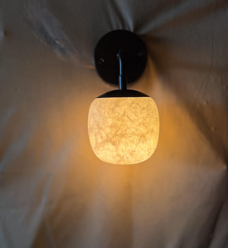 May include: A wall-mounted light fixture with a black circular base and curved arm supporting a textured, off-white globe-shaped shade. The shade emits a warm, inviting glow. The fixture is designed for indoor use.