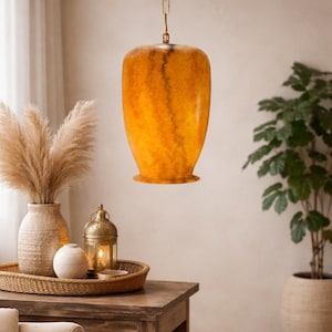 Może przedstawiać: Wisząca bursztynowa lampa wisząca ze złotym łańcuchem. Oprawa oświetleniowa ma zaokrąglony kształt przypominający wazon i teksturowaną powierzchnię. Ciepłe oświetlenie tworzy przytulną atmosferę. Światło wisi nad drewnianym stołem z elementami dekoracyjnymi.