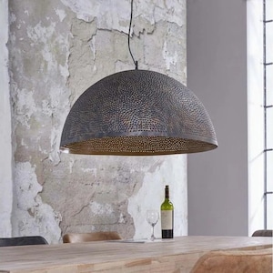 Op de afbeelding: Een grote, ronde, zwarte metalen hanglamp met een gehamerde textuur en een geperforeerd ontwerp. De lamp hangt boven een houten tafel met een glas wijn en een fles wijn op de tafel.