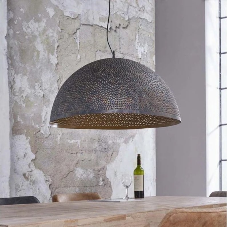 Op de afbeelding: Een grote, ronde, zwarte metalen hanglamp met een gehamerde textuur en een geperforeerd ontwerp. De lamp hangt boven een houten tafel met een glas wijn en een fles wijn op de tafel.