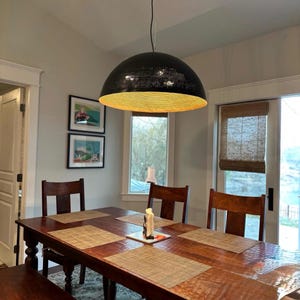 Op de afbeelding: Een zwarte metalen hanglamp met een gouden binnenkant hangt boven een houten eettafel met vier stoelen. De tafel heeft placemats en een kaarsenhouder.