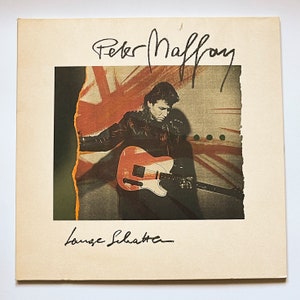 May include: A vintage record album cover featuring a musician playing a guitar. The cover is a collage of images, including a Union Jack flag and a close-up of the musician's face. The album is titled "Longue Schatten" and is signed by "Peter Maffay".