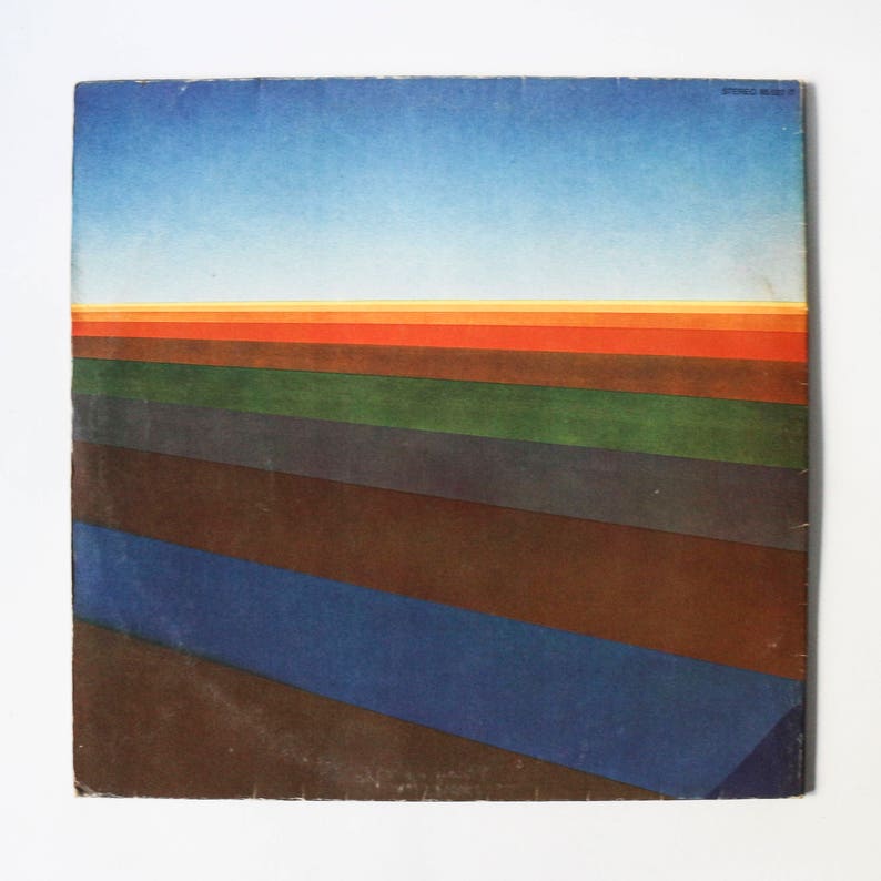Pu&ograve; includere: Una copertina di un album di vinile vintage con un design astratto colorato. La copertina presenta un cielo blu con un tramonto giallo, arancione e rosso. La met&agrave; inferiore della copertina presenta una serie di strisce orizzontali in tonalit&agrave; di marrone, verde e blu.
