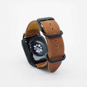 May include: A brown leather watch strap with black metal hardware. The strap is designed for a smartwatch and has a buckle fastening.