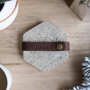 May include: A hexagonal grey felt coaster with a brown leather strap and a brass button. The strap has the word "Halpert" embossed on it.