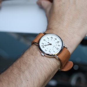 K&ouml;nnte beinhalten: Eine silberfarbene Armbanduhr mit wei&szlig;em Zifferblatt und schwarzen Ziffern. Die Uhr hat ein braunes Lederarmband. Die Uhr befindet sich am Handgelenk einer Person.