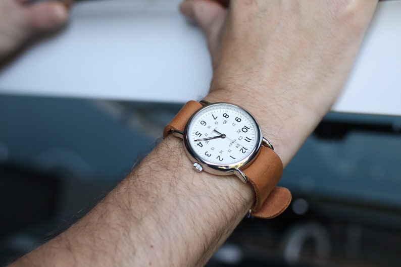 K&ouml;nnte beinhalten: Eine silberfarbene Armbanduhr mit wei&szlig;em Zifferblatt und schwarzen Ziffern. Die Uhr hat ein braunes Lederarmband. Die Uhr befindet sich am Handgelenk einer Person.