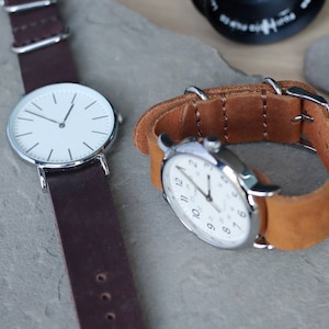 K&ouml;nnte beinhalten: Zwei Armbanduhren mit wei&szlig;en Zifferbl&auml;ttern und silberfarbenen Geh&auml;usen. Eine Uhr hat ein dunkelbraunes Lederarmband, die andere ein hellbraunes Lederarmband. Die Uhren werden auf einer grauen Steinoberfl&auml;che pr&auml;sentiert.