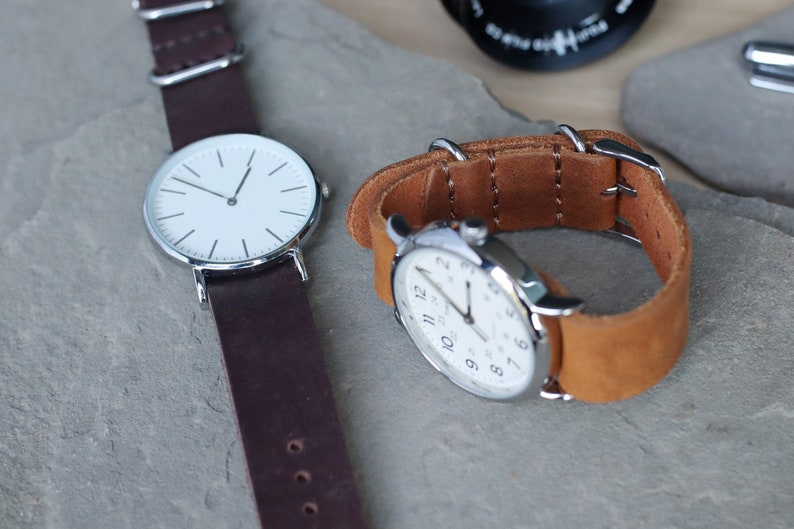 K&ouml;nnte beinhalten: Zwei Armbanduhren mit wei&szlig;en Zifferbl&auml;ttern und silberfarbenen Geh&auml;usen. Eine Uhr hat ein dunkelbraunes Lederarmband, die andere ein hellbraunes Lederarmband. Die Uhren werden auf einer grauen Steinoberfl&auml;che pr&auml;sentiert.