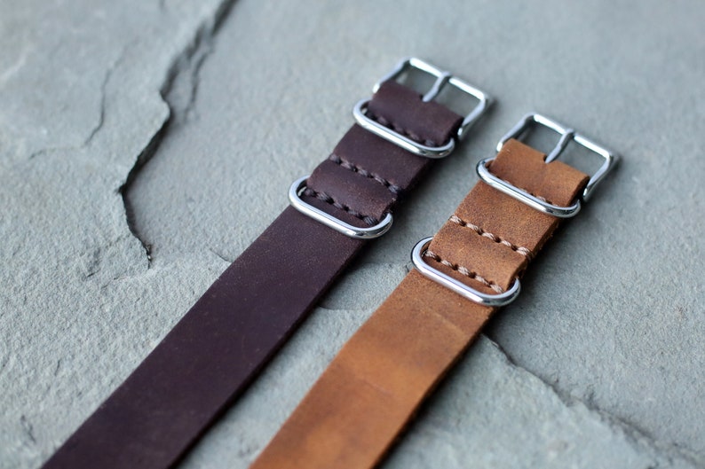 K&ouml;nnte beinhalten: Zwei Leder-Armb&auml;nder f&uuml;r Uhren, eines in Dunkelbraun und eines in hellem Braun. Beide haben silberfarbene Metallschnallen und -schlaufen. Die Armb&auml;nder sind mit einem Kontrastfaden gen&auml;ht.
