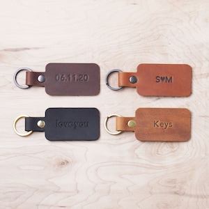 May include: Four leather keychains with different engravings. The keychains are brown, black, and tan. The engravings read "06.11.20", "S♥M", "love you", and "Keys".