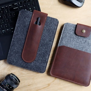 May include: Two felt and leather tablet cases. One case is gray with a brown leather pen holder. The other case is gray with a brown leather flap.