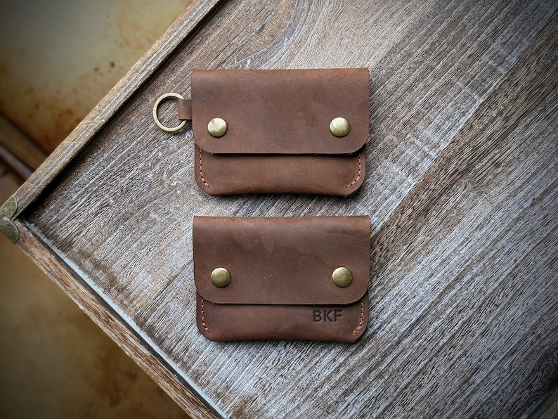 May include: Two brown leather wallets with brass accents. The top wallet has a key ring attached. The bottom wallet has the initials "BKF" embossed on the front.