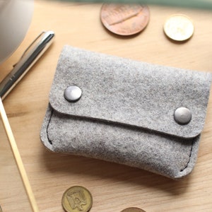 May include: A gray felt wallet with a fold-over flap and two silver snaps. The wallet is lying on a wooden surface.