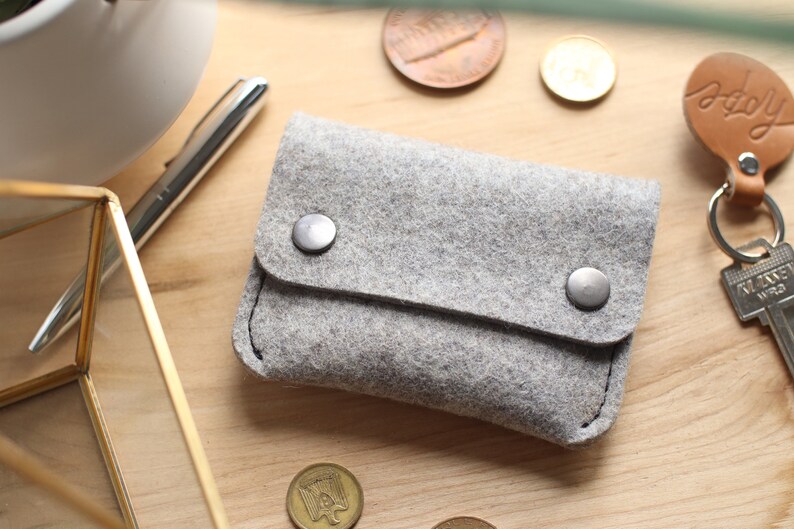 May include: A gray felt wallet with a fold-over flap and two silver snaps. The wallet is lying on a wooden surface.