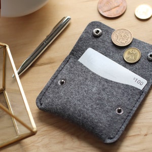 May include: A gray felt wallet with a white gift card inside. The wallet has a flap with a snap closure and several metal rivets. There are three coins on top of the flap, and the gift card has the text "$100" printed on it.