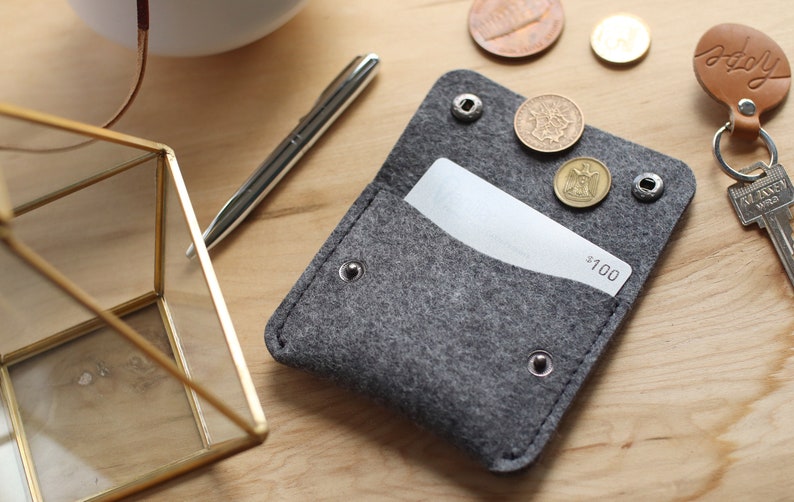 May include: A gray felt wallet with a white gift card inside. The wallet has a flap with a snap closure and several metal rivets. There are three coins on top of the flap, and the gift card has the text "$100" printed on it.