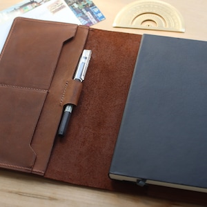 May include: A brown leather notebook cover with a pen holder and a black notebook inside.