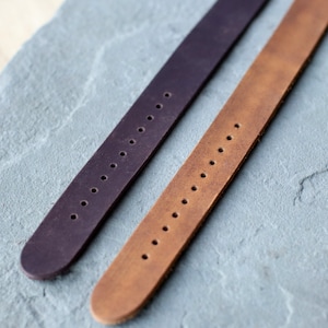 K&ouml;nnte beinhalten: Zwei Leder-Armb&auml;nder, eines in Dunkelviolett und eines in Hellbraun, werden auf einer grauen Steinoberfl&auml;che gezeigt. Jeder Riemen hat eine Reihe von gleichm&auml;&szlig;ig verteilten L&ouml;chern zur Anpassung. Die Riemen sind abgewinkelt und zeigen ihre Textur und Farbe.