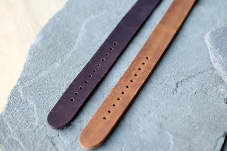 K&ouml;nnte beinhalten: Zwei Leder-Armb&auml;nder, eines in Dunkelviolett und eines in Hellbraun, werden auf einer grauen Steinoberfl&auml;che gezeigt. Jeder Riemen hat eine Reihe von gleichm&auml;&szlig;ig verteilten L&ouml;chern zur Anpassung. Die Riemen sind abgewinkelt und zeigen ihre Textur und Farbe.