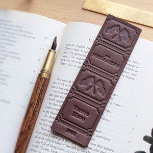 May include: A brown leather bookmark with embossed details. The bookmark features the text "Les Miserables" and "Victor Hugo" with the year "1862".