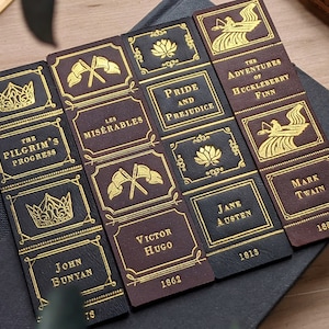 May include: Set of six leather bookmarks featuring classic literature titles. Each bookmark has a gold embossed design and title, including "The Pilgrim's Progress", "Les Misérables", "Pride and Prejudice", "The Adventures of Huckleberry Finn", and "Jane Austen".
