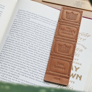May include: A brown leather bookmark with a gold embossed design. The bookmark features a crown, the text "The Pilgrim's Progress", the author's name "John Bunyan", and the year "1678".