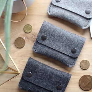 May include: Three gray felt coin purses with black snap closures. The purses are arranged in a row on a wooden surface.