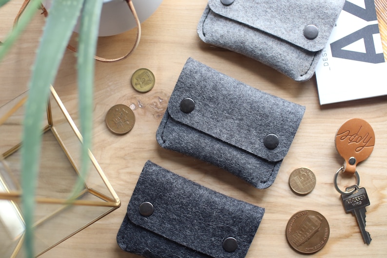 May include: Three gray felt coin purses with black snap closures. The purses are arranged in a row on a wooden surface.