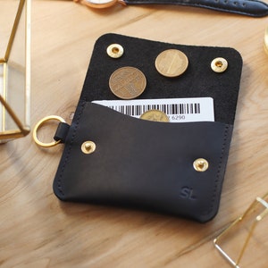 May include: A black leather wallet with a gold key ring, a small pocket for bills, and a separate compartment for coins. The wallet has gold rivets and the initials "SL" embossed on the front.