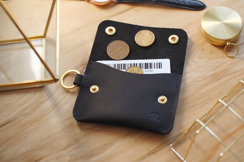 May include: A black leather wallet with a gold key ring, a small pocket for bills, and a separate compartment for coins. The wallet has gold rivets and the initials "SL" embossed on the front.