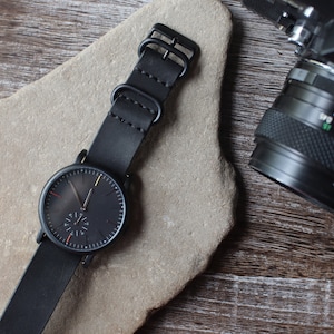 K&ouml;nnte beinhalten: Eine schwarze Armbanduhr mit Lederarmband liegt auf einer Steinfl&auml;che. Das Zifferblatt der Uhr ist dunkel mit minimalen Markierungen und einem kleinen Sekundenzeiger. Ein Kameraobjektiv ist im Hintergrund zu sehen.