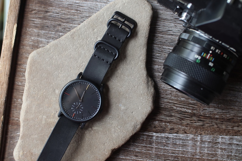 K&ouml;nnte beinhalten: Eine schwarze Armbanduhr mit Lederarmband liegt auf einer Steinfl&auml;che. Das Zifferblatt der Uhr ist dunkel mit minimalen Markierungen und einem kleinen Sekundenzeiger. Ein Kameraobjektiv ist im Hintergrund zu sehen.
