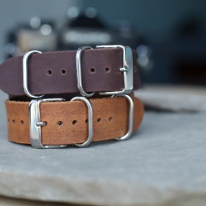 K&ouml;nnte beinhalten: Zwei Leder-Armb&auml;nder, eines in Dunkelbraun und eines in hellem Braun, &uuml;bereinander gestapelt. Beide Armb&auml;nder haben silberfarbene Schnallen und Beschl&auml;ge. Die Armb&auml;nder liegen auf einer grauen Oberfl&auml;che. Uhrenarmband.