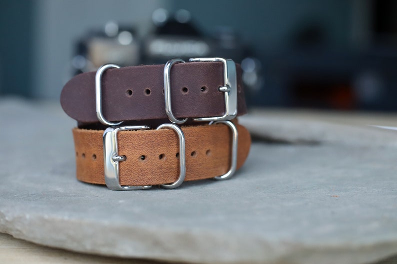 K&ouml;nnte beinhalten: Zwei Leder-Armb&auml;nder, eines in Dunkelbraun und eines in hellem Braun, &uuml;bereinander gestapelt. Beide Armb&auml;nder haben silberfarbene Schnallen und Beschl&auml;ge. Die Armb&auml;nder liegen auf einer grauen Oberfl&auml;che. Uhrenarmband.